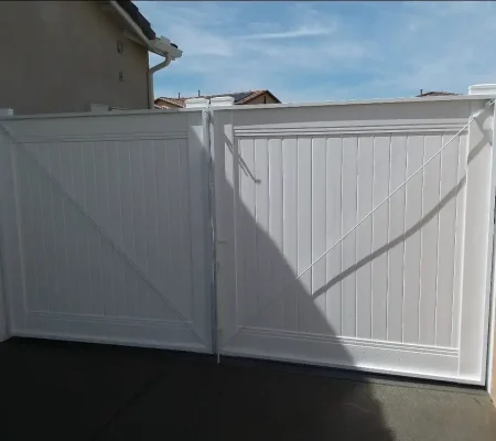 A white wooden fence with vertical slats stands in a concrete driveway in front of a beige-colored house with a closed gate.