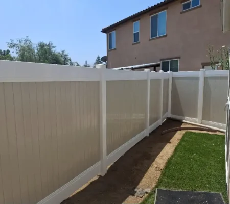 Residential yard with well-maintained green grass, wooden white fence, concrete step, small hose, and a house with brown and red tile roofs.