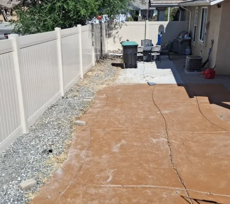 Residential construction site with a concrete patio being prepared for a new surface surrounded by a white fence and gravel.