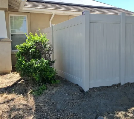 A beige house with a brown roof, a white wooden fence, and a green bush in a residential yard with a clear blue sky.