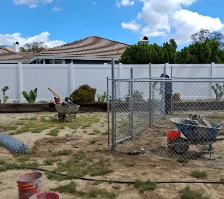 A white house with a white fence surrounded by a yard with green plants, a blue sky with white clouds, and gardening tools.