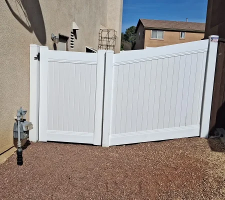 A white gate stands open in front of a beige wall, revealing a wooden fence and a fire hydrant on a gravelly ground with a house in the background.