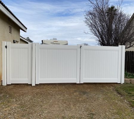 White vinyl double gate installed between two houses with a dirt driveway and a tree in the background under a partly cloudy sky.