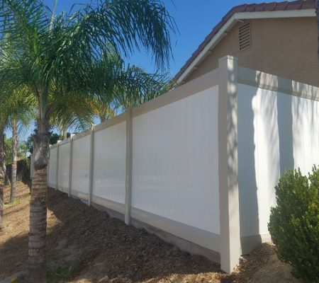 A white fence stands in front of several tall palm trees against a clear sky.