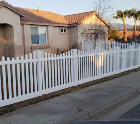 Single-story beige house with a white picket fence, palm trees, and a sidewalk along a quiet suburban street at sunset.