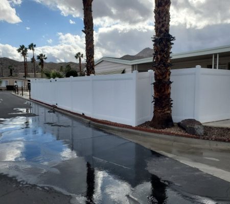 Wet street reflecting cloudy sky, white vinyl fence, palm trees, and desert mountains in a suburban neighborhood.