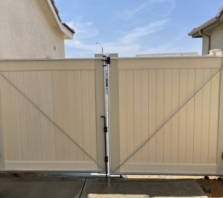 Double beige vinyl gate with metal hinges and diagonal support braces between two houses under a clear blue sky.