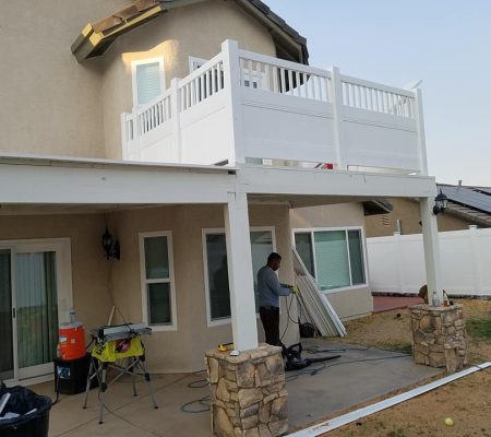 Man working on a covered patio with stone pillars and a white balcony above attached to a beige stucco house.