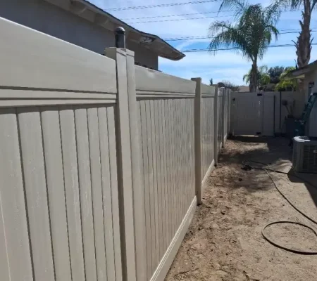 Residential area with a wooden white fence, a hose on the ground, and a hose reel near a house with a white roof and wall.