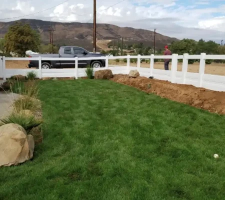 Residential area with a white picket fence, a brown-roofed house, and a well-maintained yard with a green lawn and a large rock formation.