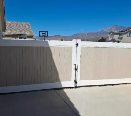 Residential area with a white fence, open to reveal a white door with a black handle, set against a clear blue sky and mountainous background.