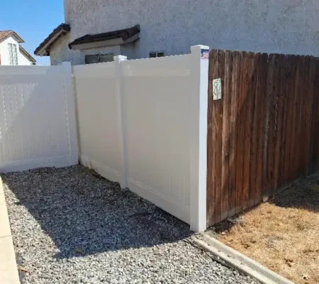 Residential scene with a white wooden fence, brown gate, and a house with a white roof and brown chimney on a gray gravel ground.