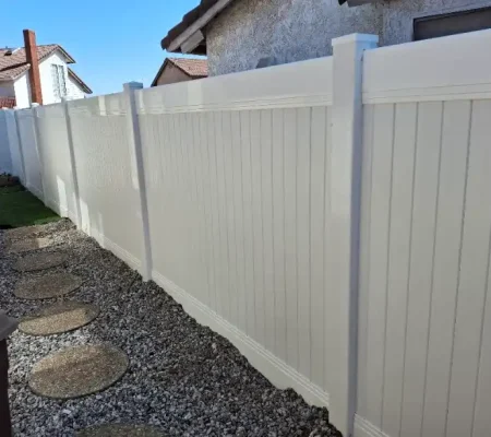 A serene residential scene featuring a white wooden fence, gravel path, stone steps, and a two-story house with a red tile roof under a clear blue sky.