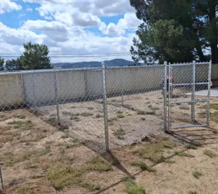 Metal chain link fence in a grassy area surrounded by a concrete wall with trees in the background under a blue sky with white clouds.