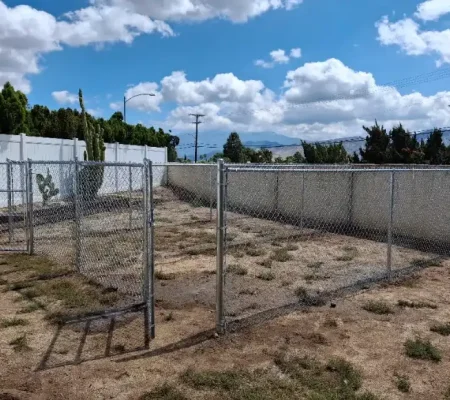 Rural yard with chain link metal fence, white and concrete walls, dry grass, trees, and a blue sky with white clouds.