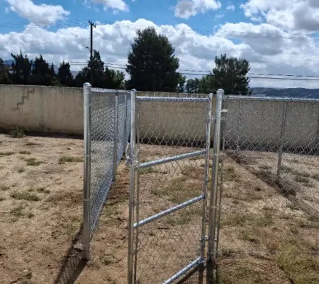 Metal chain link fence with open gate in a dry brown grassy yard surrounded by trees and telephone poles under a blue sky with white clouds.
