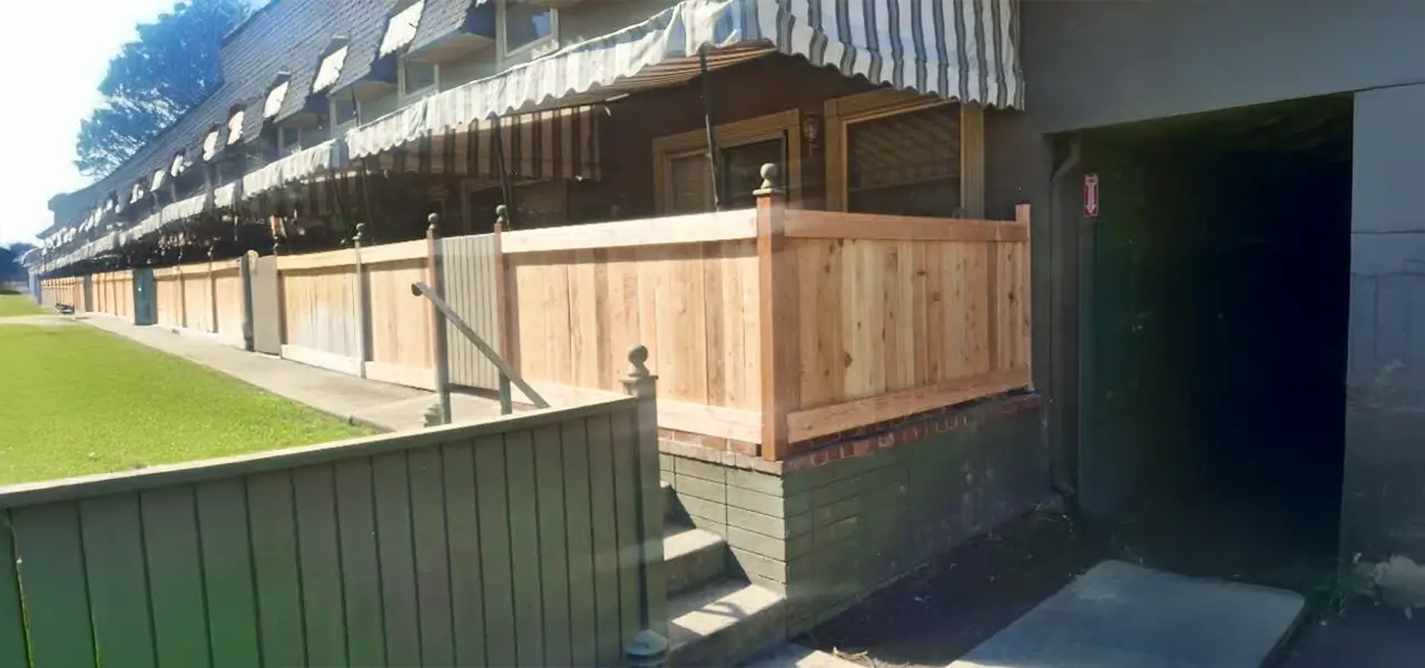 A gray house with a wooden deck and white and blue striped awning, surrounded by a wooden fence and a green bush.