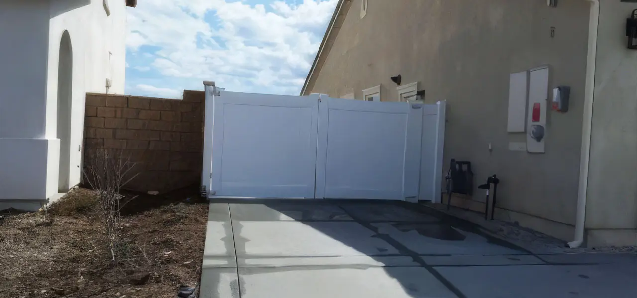 A residential area with a white gate, beige wall, and a small tree, set against a clear blue sky with fluffy white clouds.