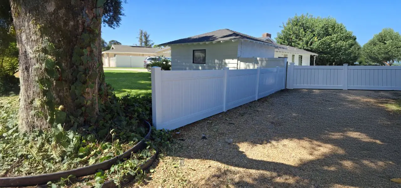 Residential scene with a white picket fence, gray-roofed house, brown tree, green leaves, gravel ground, garden hose, and a clear blue sky.