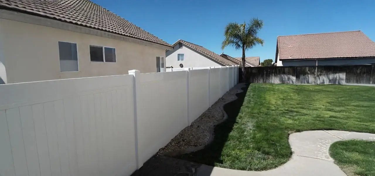 A residential area enclosed by a white fence, featuring a brown-roofed house and a tall green palm tree with a brown trunk.