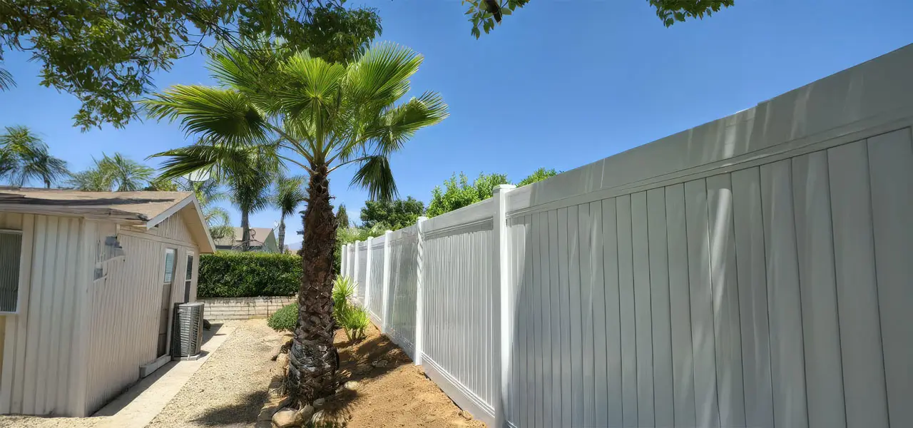 A serene residential scene featuring a white slanted fence, a palm tree with swaying green fronds, and a small white shed under a clear blue sky.